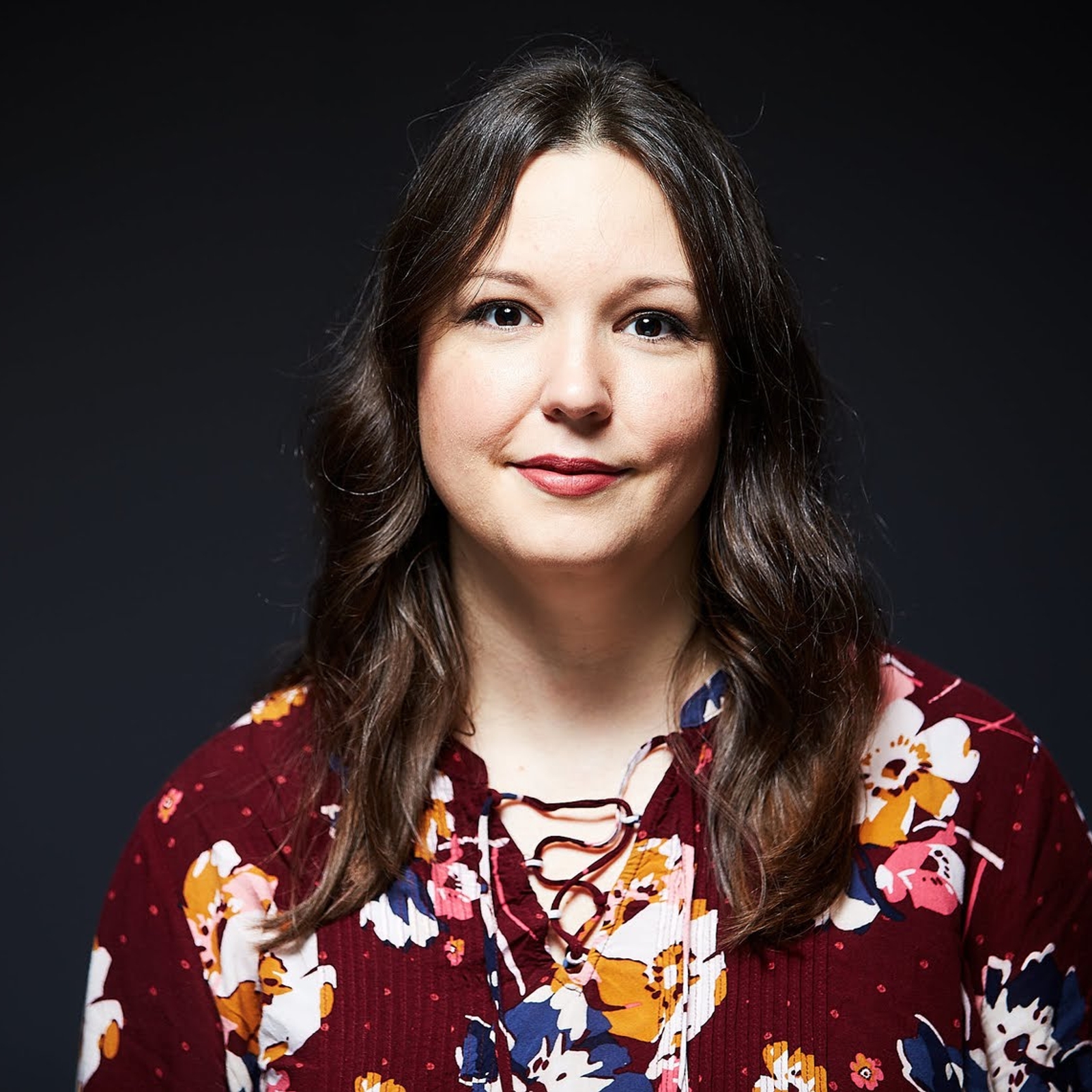 Professional head and shoulders shot of a woman in a maroon floral blouse, with long brown hair, looking directly at the camera